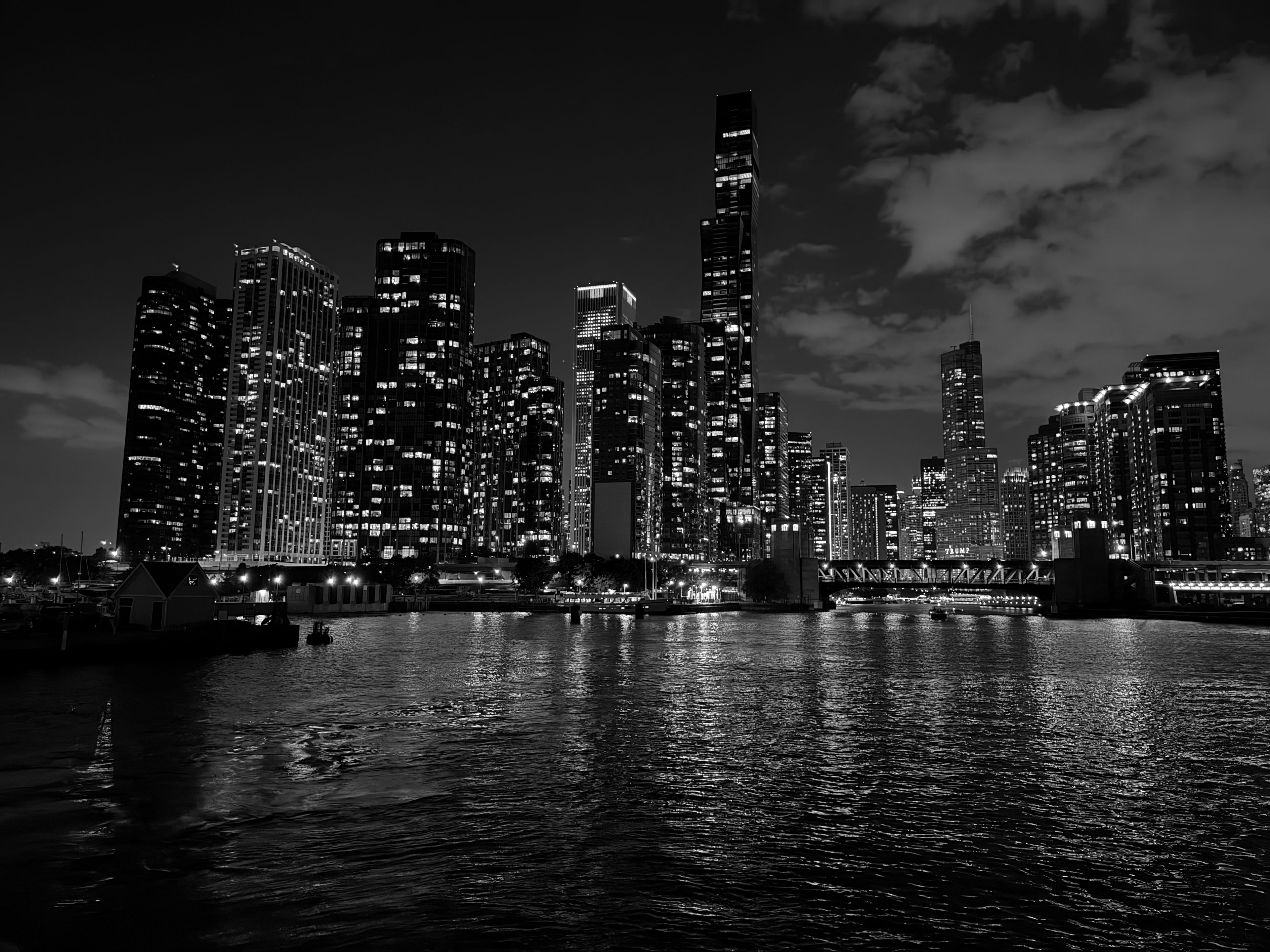 Illuminated Chicago high-rises reflected in the dark river, with a small boathouse and a low bridge along the near bank.