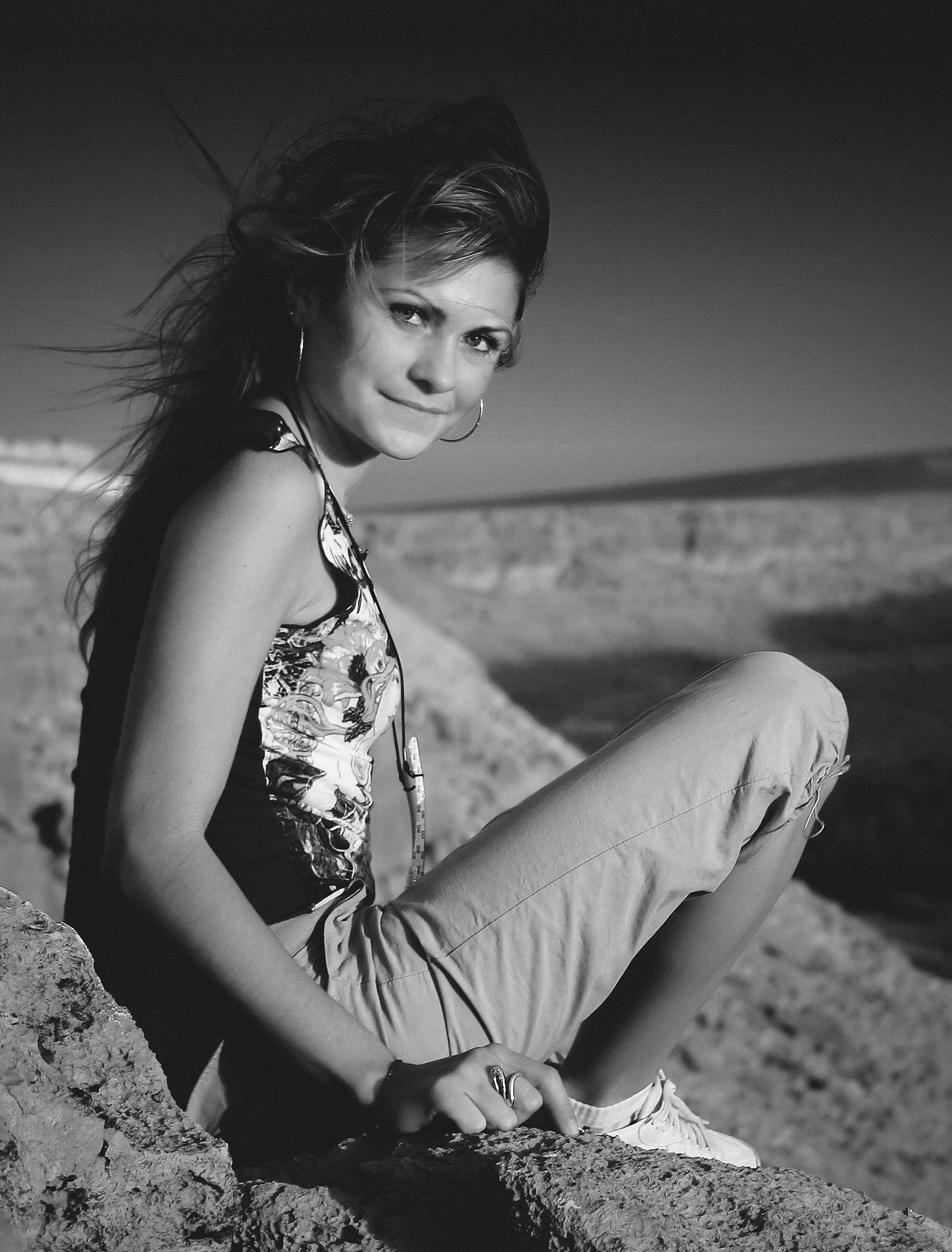 A woman sits on a rocky ledge, looking back towards the camera against a distant landscape.