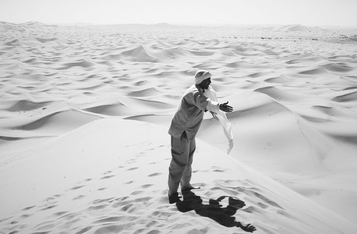A man stands on a dune holding a light scarf, with rippling sand stretching behind him.