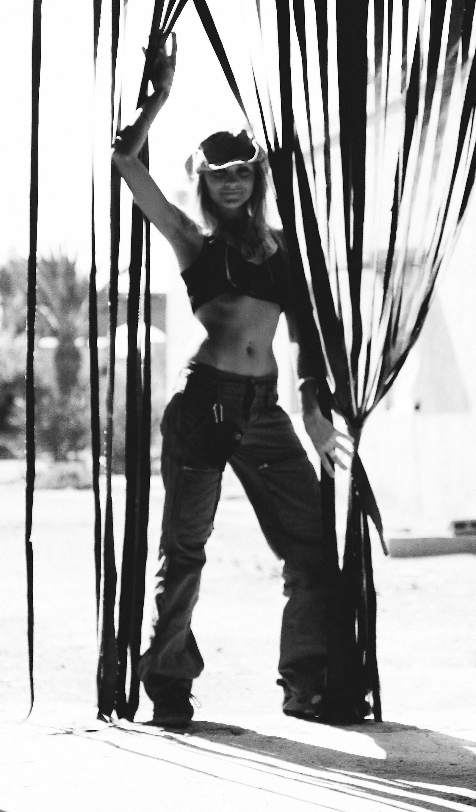 A woman stands between long hanging curtains with one arm raised against the bright light outside.