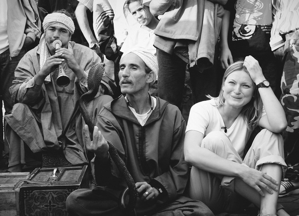 A woman sits beside two street performers, one playing a wind instrument and another handling a snake, as spectators gather behind.