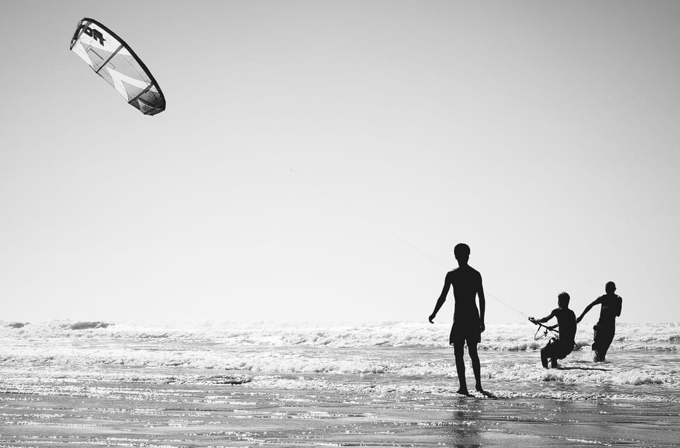 Three silhouetted figures stand in the surf beneath a kite against a bright sea and sky.