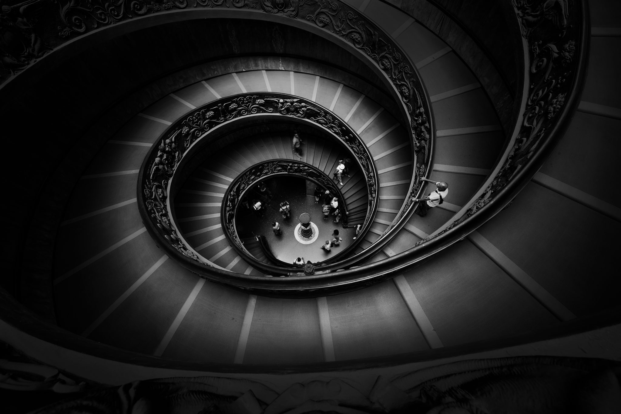 A spiral staircase curves down around a central opening, with ornate railings and small figures on the steps.
