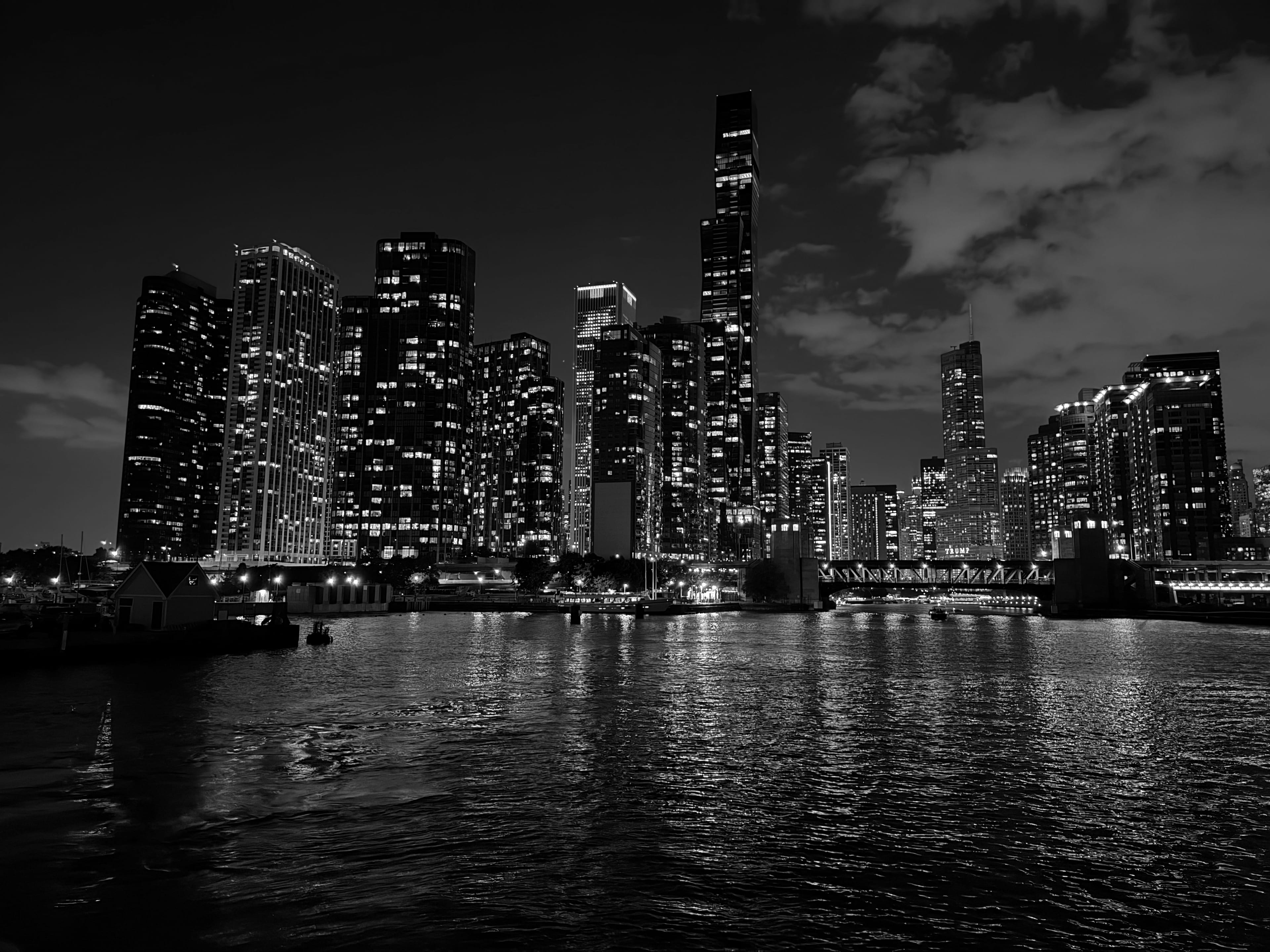 A dense cluster of illuminated high-rises reflected in the dark river at blue hour, with a small boathouse and a low bridge along the near bank.