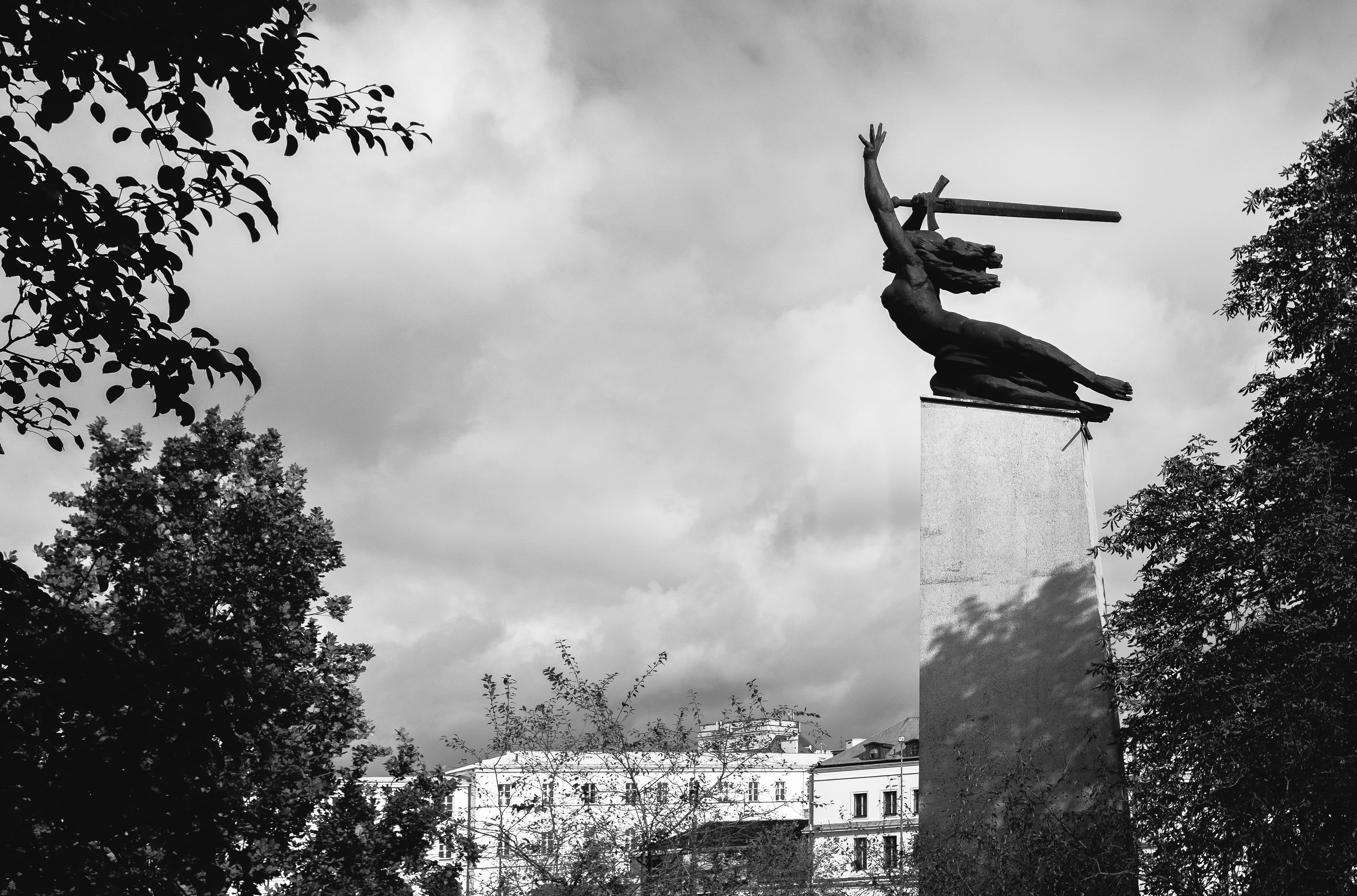 Warsaw's mermaid monument raises a sword above trees and buildings beneath a cloudy sky.